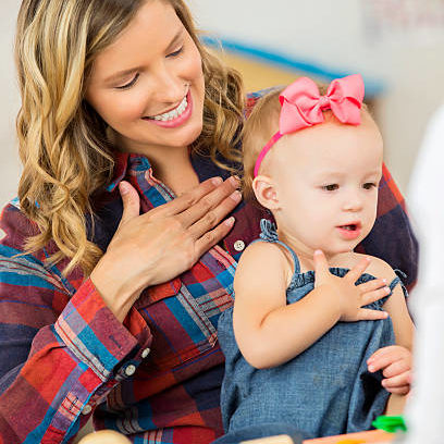 Precious little blonde toddler girl sitting on pretty Caucasian mother's lap. Mother is smiling and practicing "please" sign language with daughter.